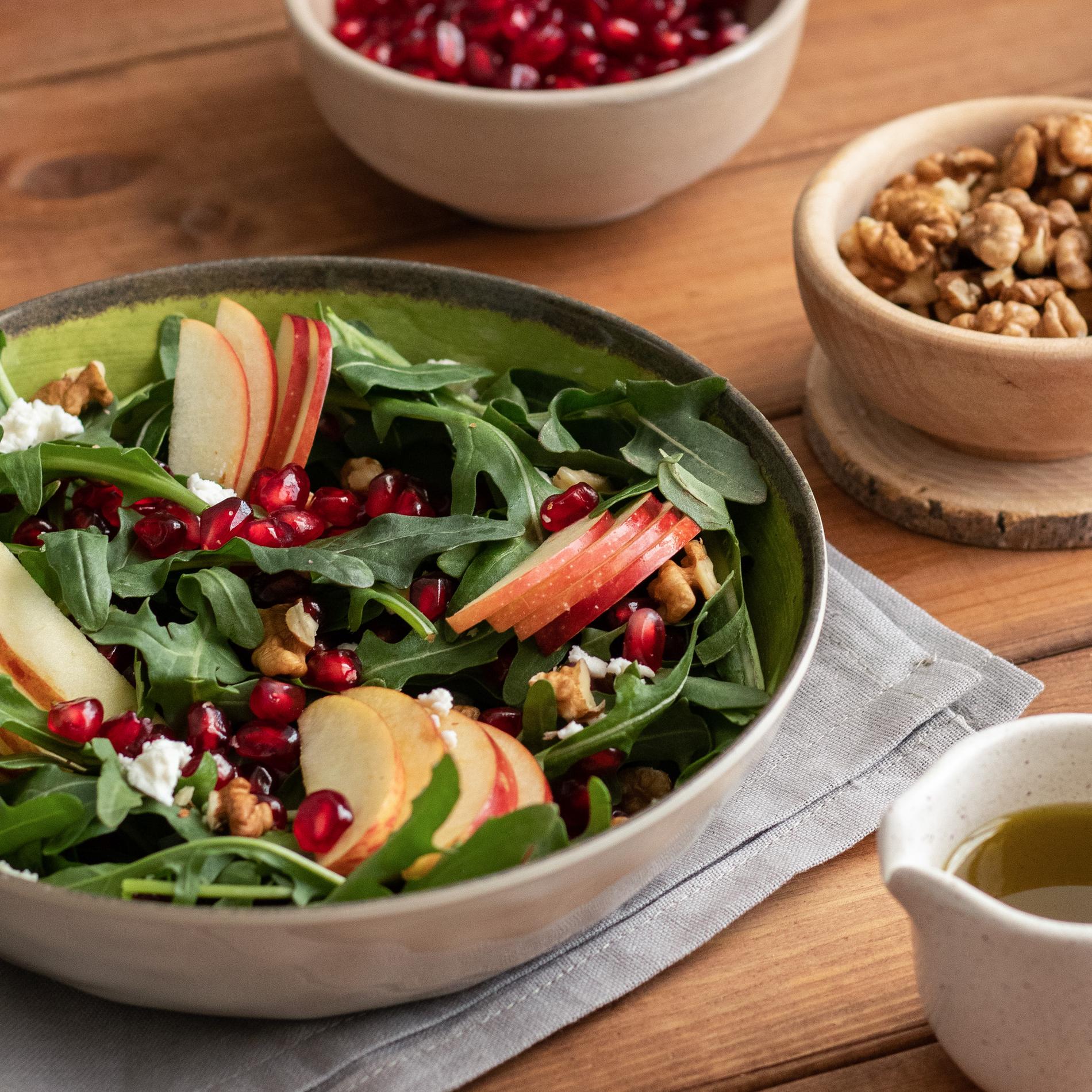 Close-up three-quarter view of a ceramic bowl filled with peppery arugula leaves, thinly sliced red apple fans, glossy pomegranate arils, crumbled white goat cheese and toasted walnut pieces on a warm wooden table; a neutral linen napkin sits beneath the bowl and a small wooden bowl of walnuts plus a pouring jug of olive oil are softly out of focus in the background. Soft natural side light and shallow depth of field emphasize the textures and jewel-like colors.
